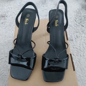 Svarta sandaler med klack från Shein - Stilrena svarta sandaler från Shein med lackad finish, fyrkantig tå och smala remmar. Snygg rosettdetalj framtill och elegant klack. Perfekta för dig som vill ha en trendig och chic look till sommaren.