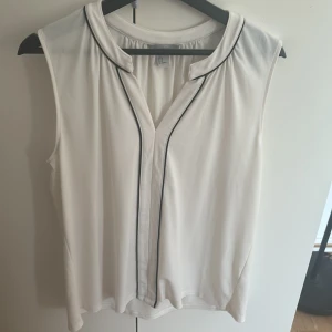 Vit ärmlös blus från H&M, M - Snygg vit ärmlös blus från H&M i storlek M. Blusen har svarta tunna kantband längs krage och knappslå, samt en lätt v-ringning. Luftigt material som känns skönt mot huden och passar perfekt till varma dagar.