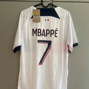 PSG Mbappé 7 vit matchtröja Nike - Snygg vit PSG fotbollströja med Mbappé 7 på ryggen. Tröjan har korta ärmar, mörkblå och röda detaljer samt Paris Saint-Germain-logga och Nike-märke framtill. Tillverkad i lätt och ventilerande material, perfekt för fotbollsplanen eller att bära till vardags.