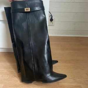 Svarta höga stövlar med spetsig tå - Snygga svarta stövlar med spetsig tå och klack. Stövlarna har en dekorativ rem med guldfärgat spänne upptill och en stilren, minimalistisk design. Perfekta för att lyfta vilken outfit som helst.