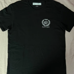 Svart T-shirt med "PHSSupport" tryck - Helt ny svart T-shirt med gotisk "PHS"-text pá ryggen och "PHS Support"-tryck pá framsidan med knogjärn och lagerkrans. Finns i alla storlekar - perfekt för dig som vill visa ditt stöd med stil. Aldrig använd,det finns alla storlekar(S,M,L,,XL