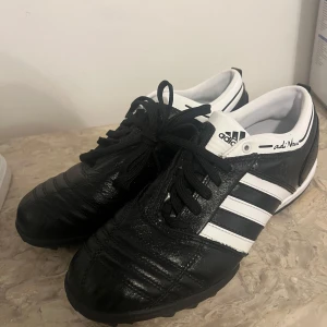 Svarta och vita Adidas adiNova fotbollsskor - Klassiska Adidas adiNova fotbollsskor i svart och vitt med de ikoniska tre ränderna på sidan. Skorna har snörning och är tillverkade i skinn för bästa känsla på planen. Perfekta för dig som vill ha stilrena och bekväma skor till fotbollsträningen.