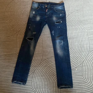 Dsquared2 blå skinny jeans med slitningar - Snygga blå jeans från Dsquared2 med skinny passform och coola slitningar samt färgstänk framtill. Klassisk femficksmodell med ikonisk Dsquared2-detalj på fickan och patch bak. Perfekta för dig som gillar streetstyle och vill sticka ut.
