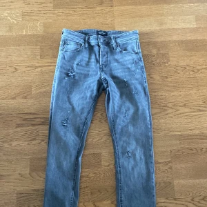 Grå slitna jeans från Jack & Jones strl w29 L32 - Snygga grå jeans från Jack & Jones med slitna detaljer och klassisk femficksmodell. Jeansen har en rak passform och är tillverkade i mjukt bomullstyg med lite stretch. Perfekta för dig som gillar en avslappnad och trendig look.