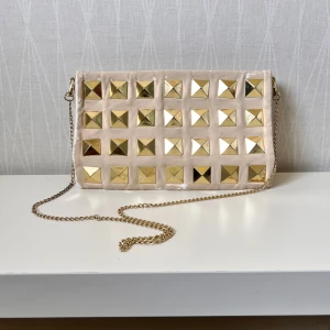 Beige kuvertväska med guldiga nitar - Kuvertväska i beige mocka med stora fyrkantiga guldnitar på framsidan och en tunn guldfärgad kedja som axelrem. Väskan har en stilren och edgy look, perfekt för att lyfta din outfit. Ingen synlig logga eller märke.