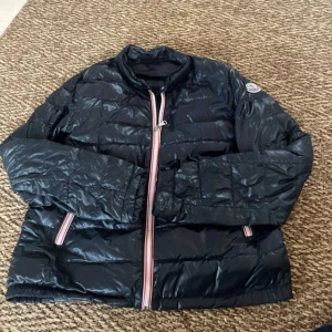 Svart Moncler jacka - Snygg svart jacka från Moncler med klassisk quiltad design och coola rödvita detaljer vid dragkedjan. Jackan har hög krage, lång ärm och Moncler-logga på ärmen. Perfekt för dig som vill ha en lätt och stilren jacka med streetkänsla.