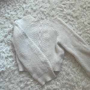 Vit stickad tröja med flätmönster - Supermysig vit stickad tröja med snyggt flätmönster framtill och på ärmarna. Tröjan har ribbade muddar vid ärmslut och midja, och är lite croppad i modellen. Perfekt för lager-på-lager under kyliga dagar.