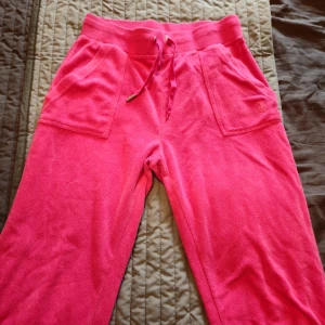 Rosa frottébyxor med snörning från Juicy Couture  - Snygga rosa mjukisbyxor från Juicy Couture i mjuk frotté. Byxorna har bred resår i midjan, snörning framtill, stora fickor fram och bak med knappar och broderad JC-logga. Perfekta för chill dagar eller när du vill vara extra comfy.