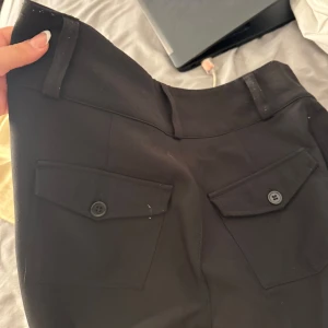 Svarta bootcut kostymbyxor med fickor - Säljer ett par svarta bootcut byxor med två stora fickor framtill med lock och knapp. Byxorna har bälteshällor och stängs med två knappar och dragkedja. Snygg passform som sitter tajt upptill och släpper ut vid benen.