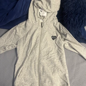Grå hoodie från Comme des Garçons Play - Grå hoodie med dragkedja från Comme des Garçons Play. Ikoniskt svart hjärta med ögon på bröstet och stort tryck på ryggen. Mjuk bomull, huva och fickor framtill. Perfekt för en chill och stilren look.