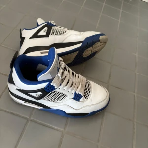Nike Air Jordan 4 Retro Motorsports - Nike Air Jordan 4 Retro sneakers i vitt och blått med svarta detaljer. Skorna har meshpaneler, snörning och den klassiska Jumpman-loggan på hälen. Ovandel i skinn och mesh, vadderad plös och krage för extra komfort. Perfekta för dig som gillar streetstyle.