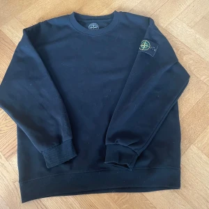 Svart sweatshirt från Stone Island - Svart sweatshirt från Stone Island med klassisk rund hals och ribbade muddar. Ikonisk logotyp-patch på vänster ärm. Tröjan är i mjuk bomull och har en clean, stilren look som passar perfekt till streetwear eller chill vardagsstil.