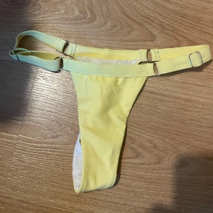Gul bikiniunderdel med justerbara band - Snygg gul bikiniunderdel med högt skuren tanga-modell och justerbara band i sidorna med metallspännen. Perfekt för dig som vill ha en trendig och minimalistisk look på stranden eller vid poolen.