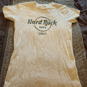 Vit Hard Rock Cafe Oslo t-shirt - Vit t-shirt från Hard Rock Cafe Oslo med glittrigt svart tryck på bröstet. Klassisk passform med rund hals och korta ärmar. Tillverkad i 100% bomull, perfekt för dig som gillar ikoniska bandtröjor och vill sticka ut lite extra.