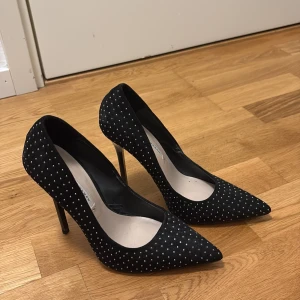 Svarta pumps med silverprickar Zara - Snygga svarta pumps från Zara Woman med spetsig tå och hög klack. Skorna är täckta av små silverfärgade prickar som ger en glittrig känsla. Ytan ser ut att vara i mocka och de har en elegant, smal siluett. Perfekta för att lyfta din outfit.