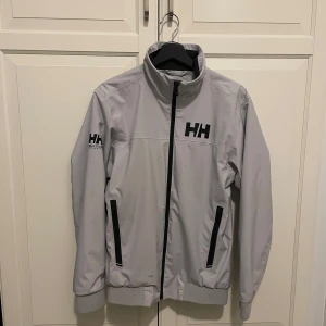Helly Hansen S Windbraker - Snygg ljusgrå vindjacka från Helly Hansen med svart HH-logga på bröstet och ärmen. Jackan har hög krage, dragkedja framtill och två svarta fickor med dragkedja. Tillverkad i slitstark polyester och perfekt för blåsiga dagar.