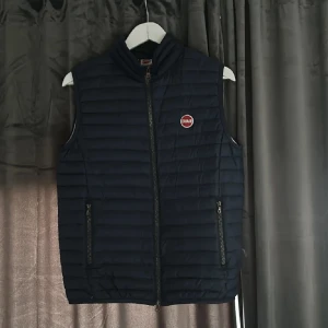 Comar väst - Snygg mörkblå dunväst från Colmar med quiltad design och dragkedja framtill. Västen har två fickor med dragkedja och en rund Colmar-logga på bröstet. Perfekt att slänga över en hoodie för en chill look.