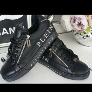 Svarta sneakers Philipp Plein med dragkedja - Svarta sneakers från Philipp Plein i skinn med coola silvriga dragkedjor och rakbladshänge. Snygg logga längs sidan och detaljer i metall. Platt sula och snörning framtill ger en edgy vibe. Perfekta för dig som vill sticka ut.