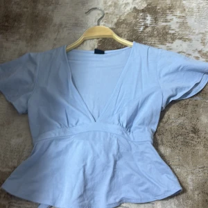 Ljusblå omlottopp från Gina Tricot - Supersöt ljusblå omlottopp från Gina Tricot i mjuk bomull. Toppen har v-ringning, korta ärmar och knytband i midjan som man knyter där bak. Storlek xs