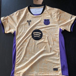 Barcelona bortatröja Nike L - Snygg FC Barcelona bortatröja från Nike i beige med lila detaljer och svart krage. Tröjan har korta ärmar, gulprickigt mönster och tryck från Spotify och UNHCR. Tillverkad i lätt Dri-FIT-material som andas, perfekt för match eller träning.
