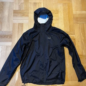 Svart vindjacka från Helly Hansen - Svart vindjacka från Helly Hansen med huva och dragkedja framtill. Jackan har två sidofickor med dragkedja och justerbar nederkant. Tillverkad i lätt och slitstark polyester, perfekt för blåsiga dagar. Klassisk och stilren design med HH-logga på bröstet.