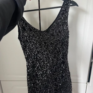 Svart glittrig paljettklänning - Supersnygg svart kortklänning täckt av glittriga paljetter. Ärmlös modell med djupare ringning fram och smal passform som sitter tajt. Perfekt för dig som vill sticka ut och glänsa på festen!