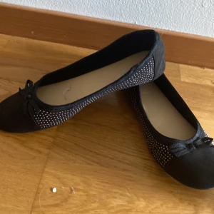 Svarta ballerinaskor med rosett och nitar - Snygga svarta ballerinaskor från Alley med små silvriga nitar längs sidorna och en söt rosett framtill. Skorna har rund tå och platt sula, samt en mjuk insida för extra komfort. Perfekta för dig som gillar en stilren men ändå glittrig look.