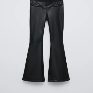 Svarta bootcut byxor i skinnimitation - Snygga svarta byxor i skinnimitation med bootcut-modell. Byxorna har en hög midja och två dekorativa fickor bak. Perfekta för dig som vill ha en edgy och trendig look.
