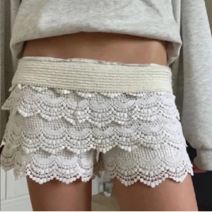 Vita virkade spets-shorts - Superfina vita shorts med bred resår i midjan och flera lager virkad spets med volangkant. De har en romantisk och bohemisk vibe, perfekta för varma dagar. Sitter skönt och har en snygg passform som framhäver benen.