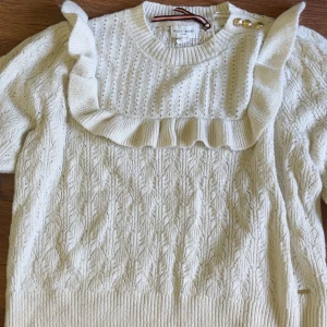 Vit stickad tröja med volang Holly & White - Supersöt vit stickad tröja från Holly & White med volangdetaljer över axlar och bröst, samt dekorativa guldknappar vid axeln. Tröjan har ribbade muddar och ett fint hålmönster upptill. Strl XL passar M