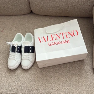 Valentino Garavani Open sneakers vit/blå - Snygga Valentino Garavani Open sneakers i vitt skinn med bred marinblå rem över sidan och coola nitar på hälen. Klassisk rund tå, platt sula och vita snören. Perfekta för dig som vill ha en lyxig och stilren look.