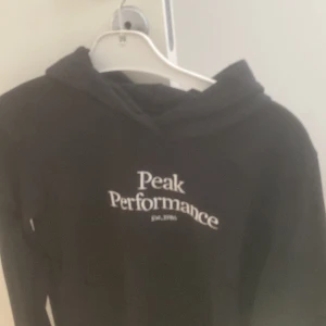 Svart hoodie från Peak Performance - Svart hoodie från Peak Performance med vit logga tryckt på bröstet. Klassisk modell med huva och långa ärmar, perfekt för en chill och sportig look. Mjuk och skön, passar till allt från jeans till träningsbyxor.