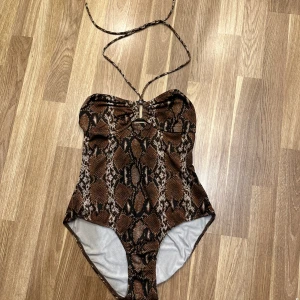 Ormmönstrad baddräkt - En ormmönstrad baddräkt i bruna och svarta toner från Esmara. Den har en snygg halterneck-design med knytband och en liten detalj framtill. Perfekt för sommarens alla badäventyr! Storlek 38