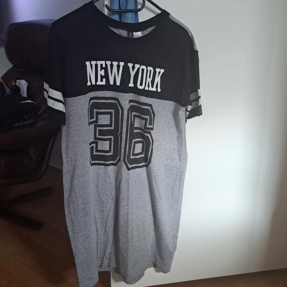 Säljer en cool t-shirt i grått och svart med texten 'New York' och siffran '36' tryckt på framsidan. T-shirten har korta ärmar med vita ränder och är perfekt för en avslappnad stil. Passar bra till jeans eller shorts för en sportig look.. T-shirts.