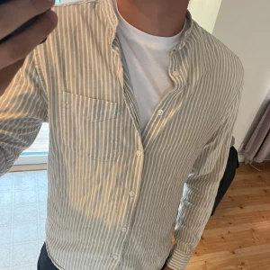 Randig skjorta från East West - Säljer en stilren randig skjorta från East West i storlek M. Skjortan är slim fit och har en klassisk design med knappar framtill och en bröstficka.  Färgerna är neutrala vilket gör den lätt att matcha med andra plagg. Mitt pris är 129kr