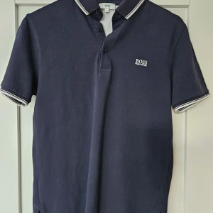 Mörkblå pikétröja från Hugo Boss - Säljer en stilren mörkblå pikétröja från Hugo Boss. Den har en klassisk krage med knappar och vita detaljer på krage och ärmslut. Perfekt för både vardag och lite finare tillfällen. Tröjan är i bra skick och redo för en ny ägare!