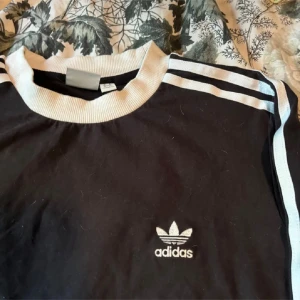 Svart långärmad tröja från Adidas - Säljer en klassisk svart långärmad tröja från Adidas med de ikoniska tre ränderna i vitt längs ärmarna. Tröjan har en vit ribbad krage och muddar, vilket ger en sportig look. Perfekt för både vardag och träning!