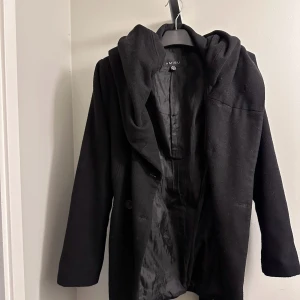 Svart kappa med huva och bälte - Snygg svart kappa från New Yorker med huva och knappar framtill. Den har ett bälte i midjan för en fin passform och långa ärmar. Perfekt för höst och vinter! Storlek 42