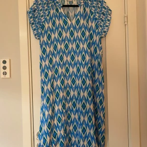 Blå, vit och grön mönstrad klänning i viskos och silke i strl M från Designed by 2-biz. - Blå, vit och grön mönstrad klänning i viskos och silke i strl M från Designed by 2-biz.  Sparsamt använd och i fint begagnat skick.  Mått Över bröstet: ca 53 cm Längd från axeln och ner: ca 105 cm  70% viskos och 30% silke 