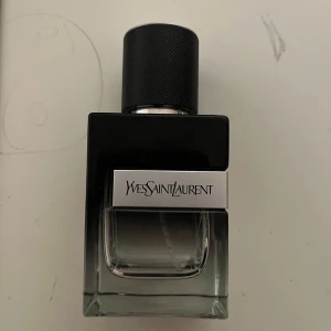 Parfym från Yves Saint Laurent - Elegant parfymflaska från Yves Saint Laurent med en stilren design. Flaskan är svart med en silverfärgad etikett och har en modern, fyrkantig form. Perfekt för den som söker en sofistikerad doft.