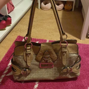 Beige handväska och plånbok från Guess - Snygg beige handväska och matchande plånbok från Guess med logotypmönster. Väskan har dubbla handtag och flera fack med dragkedja. Plånboken har flera kortfack och en transparent ID-ficka. Tillverknings år 2003