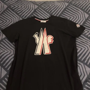 Svart t-shirt från Moncler - Snygg svart t-shirt från Moncler med ett stort, broderat logotypmotiv i vitt och rött på framsidan. T-shirten har korta ärmar och en liten Moncler-logga på ärmen. Perfekt för en stilren look. Skriv till mig vid minsta fundering! Köpt i somras aldrig använd. Priset går att diskutera.