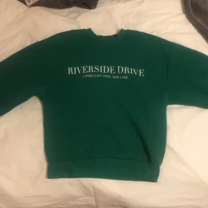 Grön sweatshirt från Gina Tricot - Säljer en grön sweatshirt från Gina Tricot med texten 'Riverside Drive, Upper East Side, New York' på framsidan. Tröjan har långa ärmar och ribbade muddar vid ärmslut och nederkant. Perfekt för en avslappnad stil!