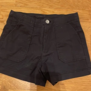 Svarta shorts från Kappahl - Snygga svarta shorts från Kappahl med hög midja och knappstängning. De har praktiska fickor både fram och bak, vilket gör dem både stiliga och funktionella. Perfekta för varma sommardagar!