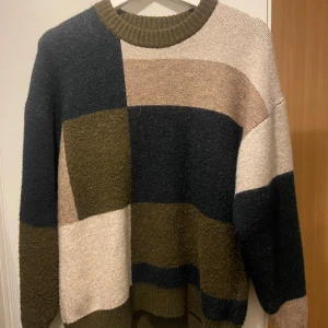 Flerfärgad stickad tröja - Säljer en mysig stickad tröja i flera färger som olivgrön, beige och mörkblå. Tröjan har ett blockmönster och långa ärmar med ribbad halsringning. Perfekt för kyliga dagar!
