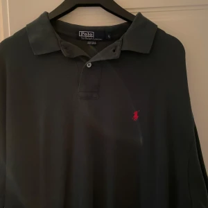 Mörkgrön  pikétröja från Ralph Lauren - Klassisk svart pikétröja från Ralph Lauren med den ikoniska röda logotypen på bröstet. Tröjan har en krage och knappar framtill, perfekt för en stilren look.