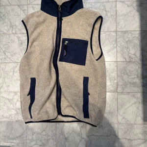 Beige fleeceväst från Patagonia - Säljer en beige fleeceväst från Patagonia med marinblå detaljer. Västen har en dragkedja framtill och en praktisk bröstficka med dragkedja. Perfekt för lager-på-lager under kyliga dagar.