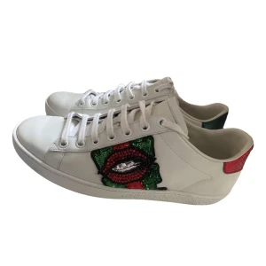  Gucci Ace  - Snygga vita sneakers från Gucci med unikt paljettbroderi i form av läppar på sidan. Skorna har snörning och detaljer i rött och grönt på hälen. Perfekta för att ge din outfit en lyxig touch.