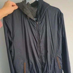 Blå och grå vindjacka med huva - Snygg och praktisk vindjacka i blått och grått med huva. Jackan har dragkedja och är perfekt för blåsiga dagar. Den är långärmad och har en stilren design.