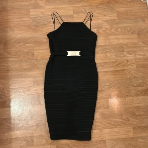 Svart klänning från Lipsy London - Elegant svart klänning från Lipsy London med smala axelband och en stilren siluett. Klänningen har en dekorativ metallspänne i midjan och en subtil ribbad textur. Perfekt för en chic och sofistikerad look. Inga fläckar eller slitage. Skriv gärna om ni har frågor.